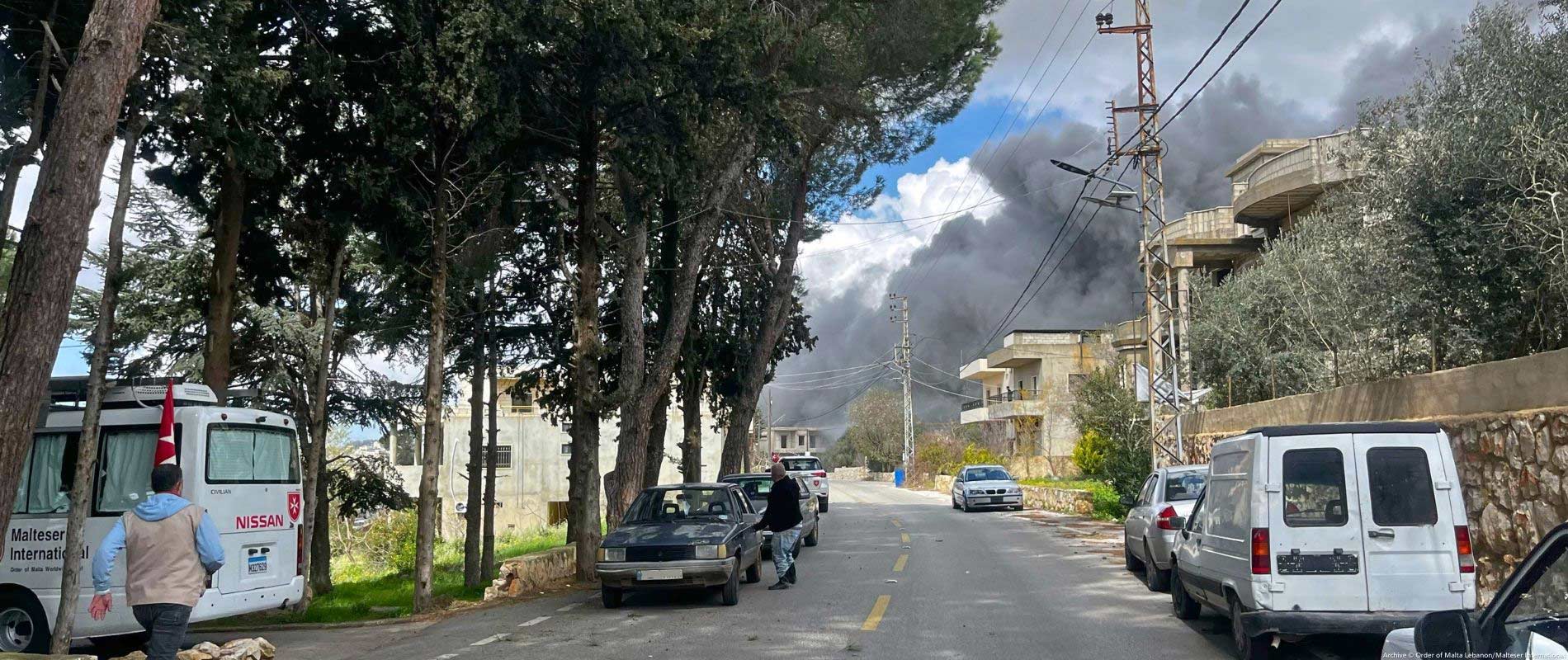 A street with cars. In the background, large clouds of smoke rise into the sky.